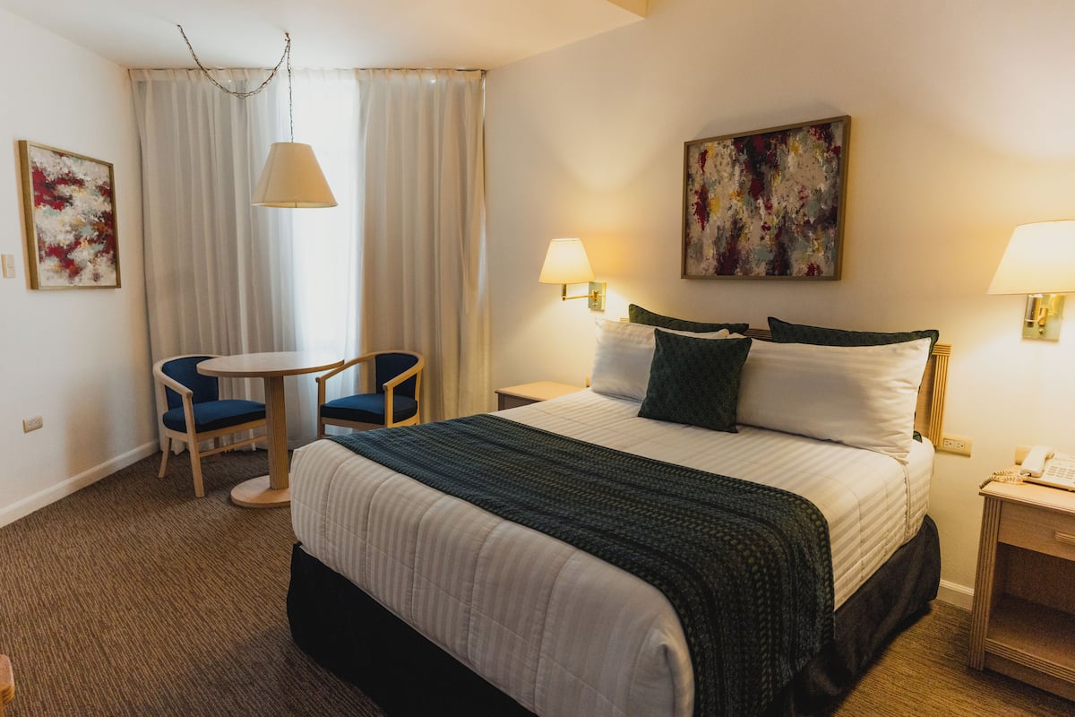 A hotel room with a bed, two bedside tables, a chair, a small table, and a window with curtains.