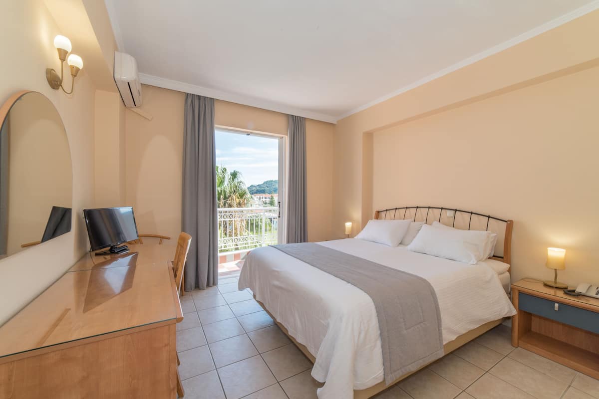 Habitación de hotel con cama, escritorio con computadora, silla, mesita de noche y balcón con vistas al exterior.