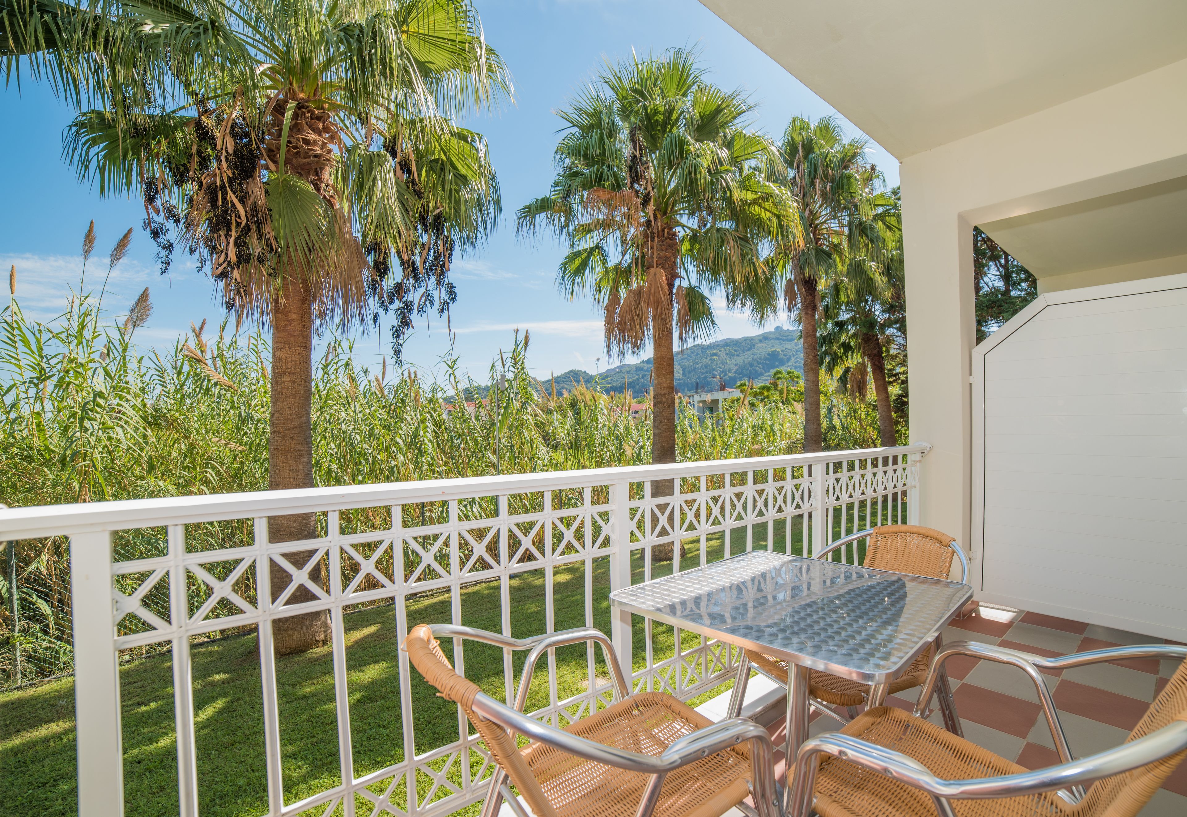 A balcony with a table and chairs, surrounded by a white lattice fence, overlooking a lush garden and palm trees.