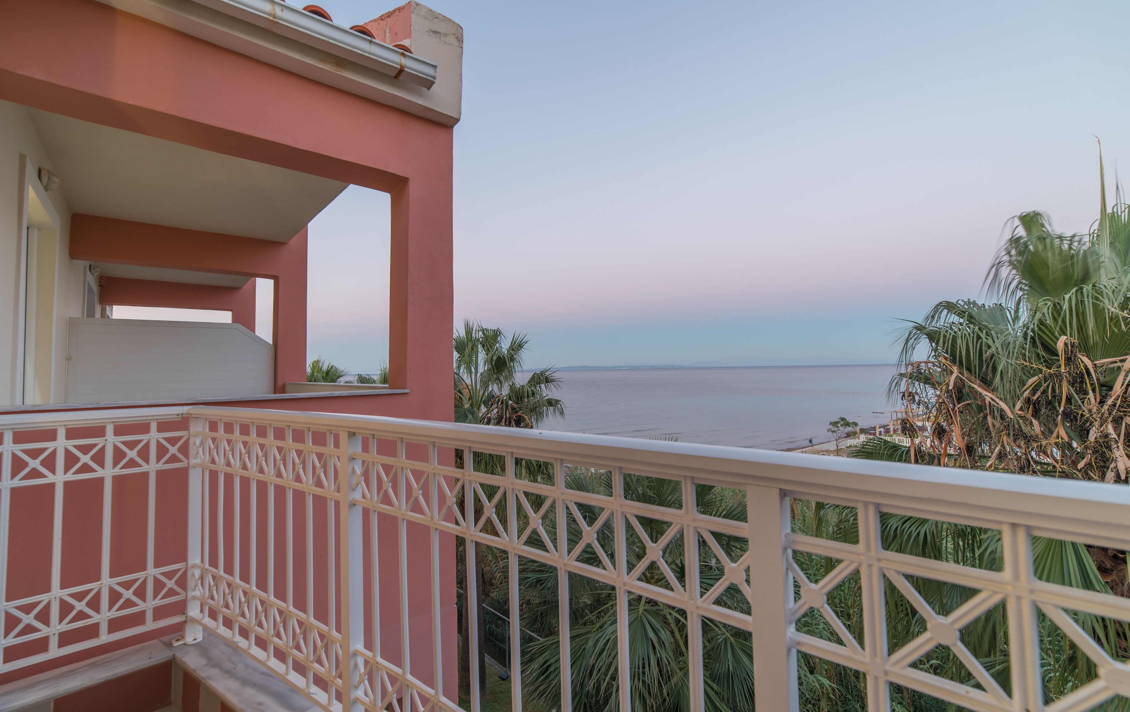 A balcony with a view of the sea, a pink building, and some palm trees.