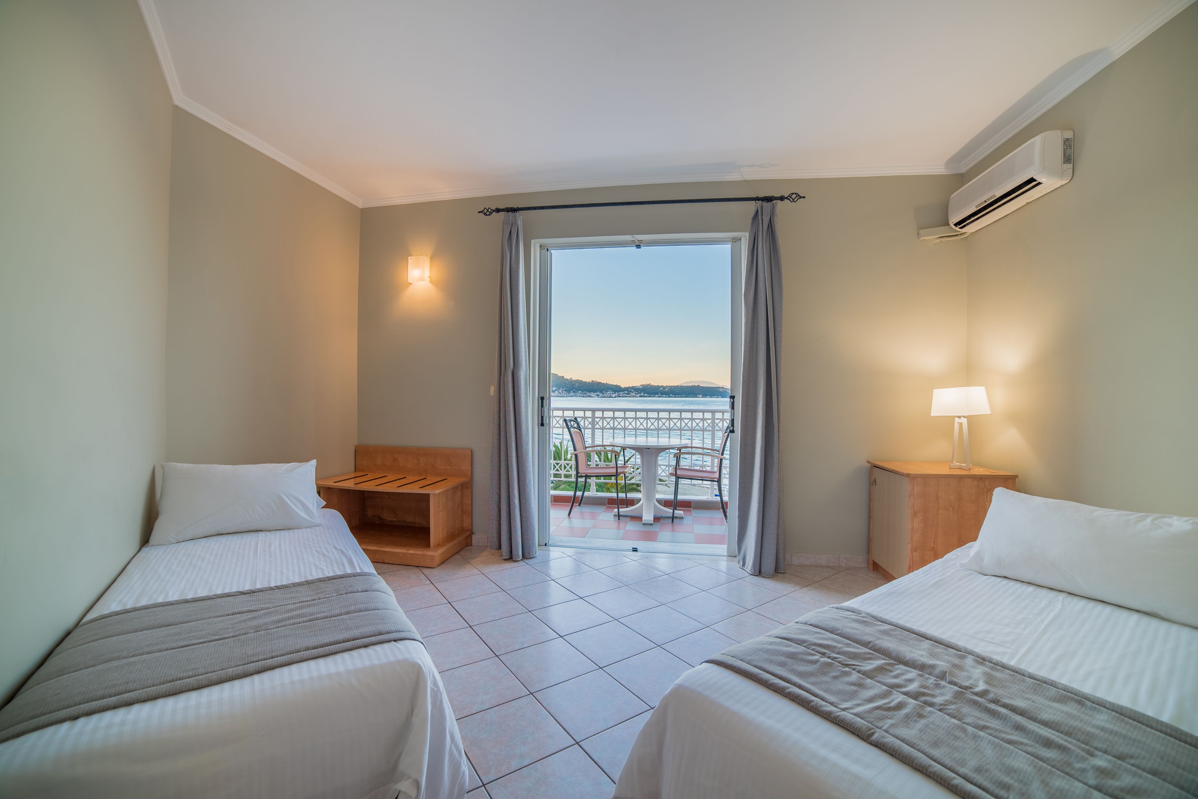 Habitación de hotel con dos camas, un balcón con vistas, un banco de madera y una mesa con sillas.