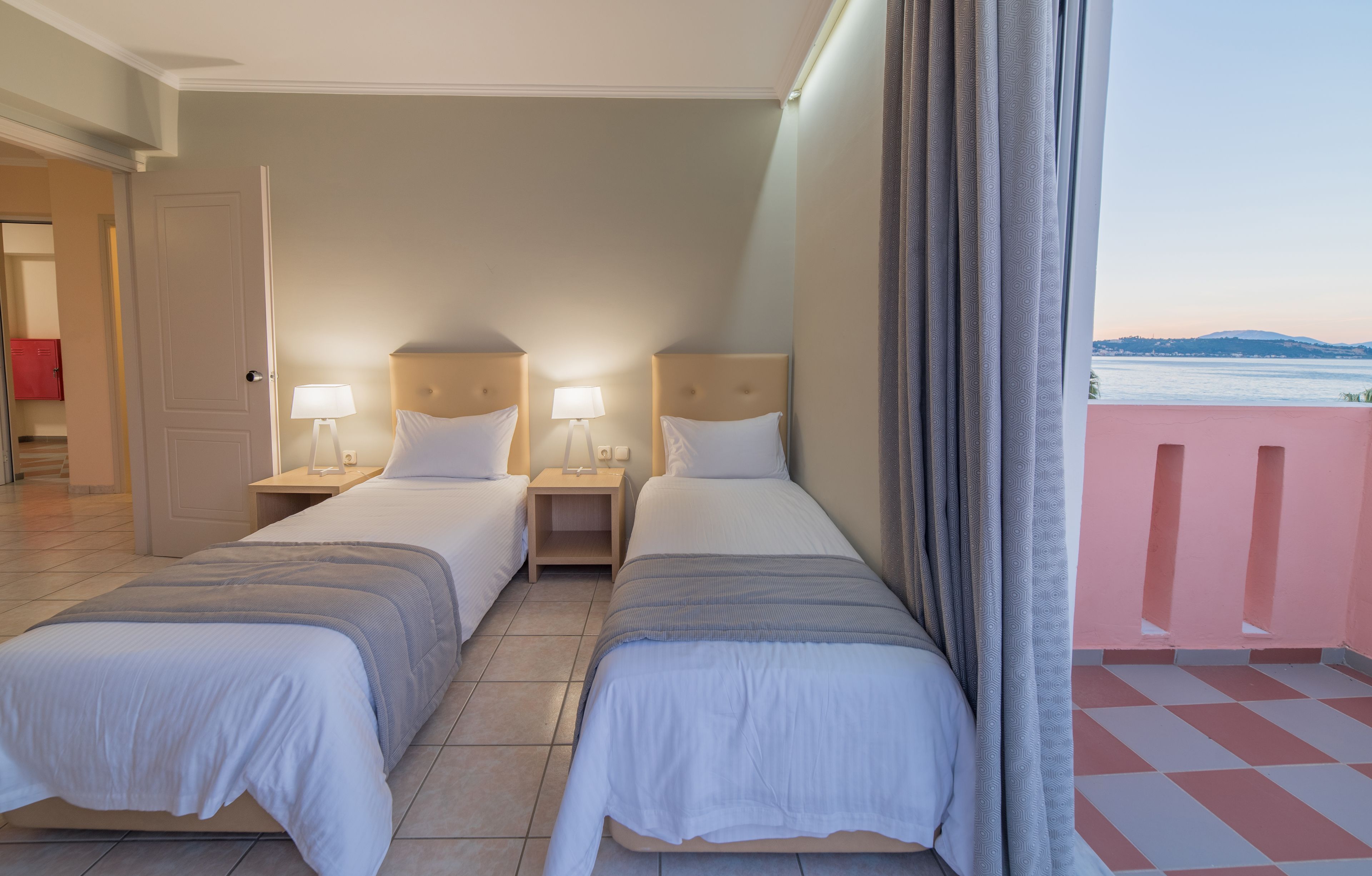 A hotel room with two beds, bedside tables with lamps, a view of the sea, and a balcony with a checkered floor.