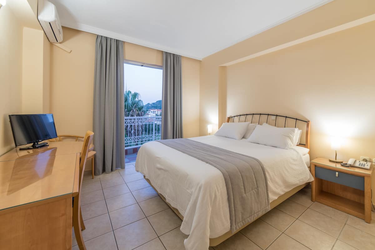 Una habitación de hotel con cama, escritorio, silla, mesita de noche y balcón con cortinas.