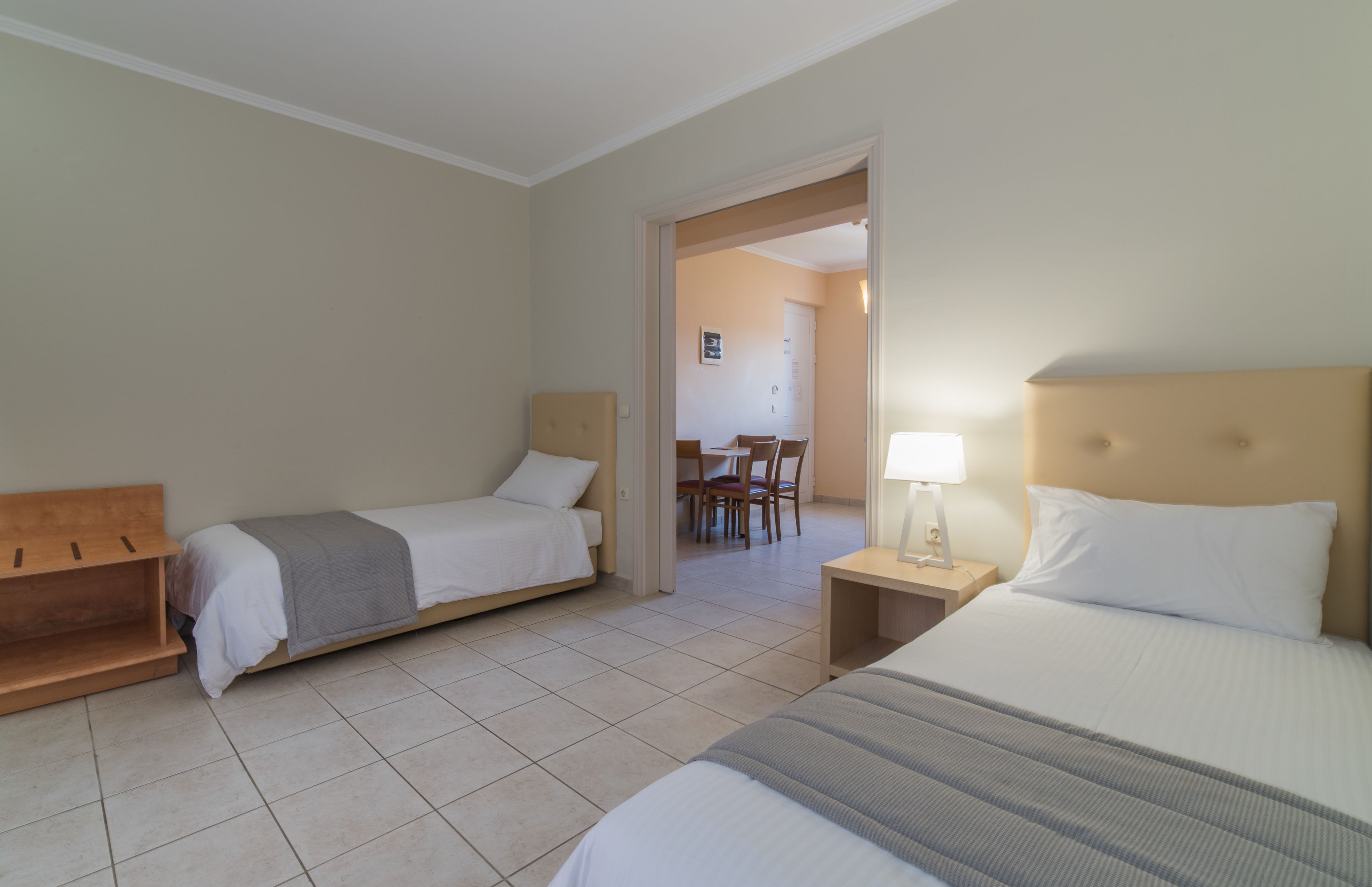 A hotel room with two single beds, a wooden bench, a desk with a lamp, and a dining area visible through an open door.