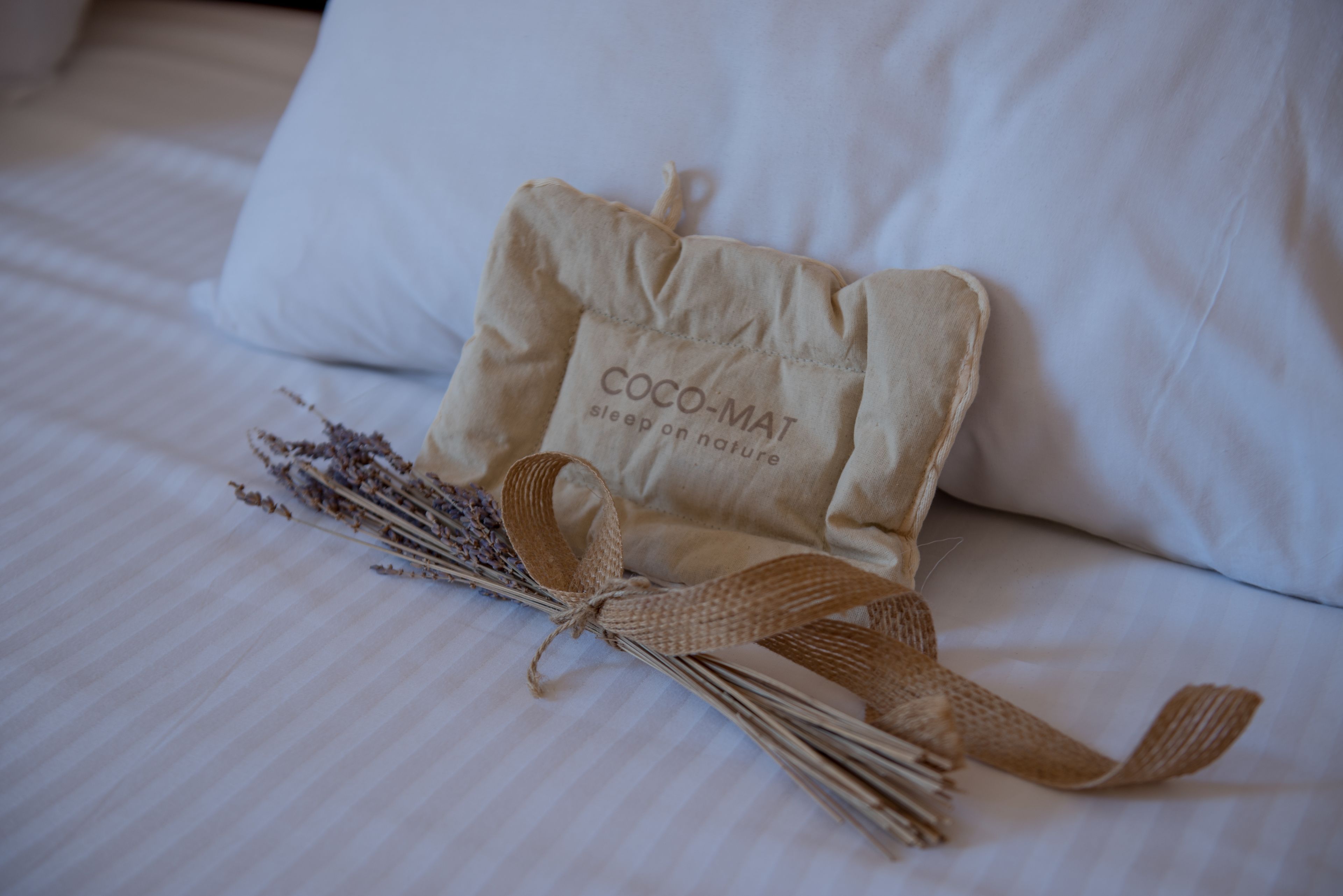 Una cama bien tendida con un almohadón blanco y una pequeña bolsa color beige con la etiqueta 'COCO-MAT sleep on nature'.