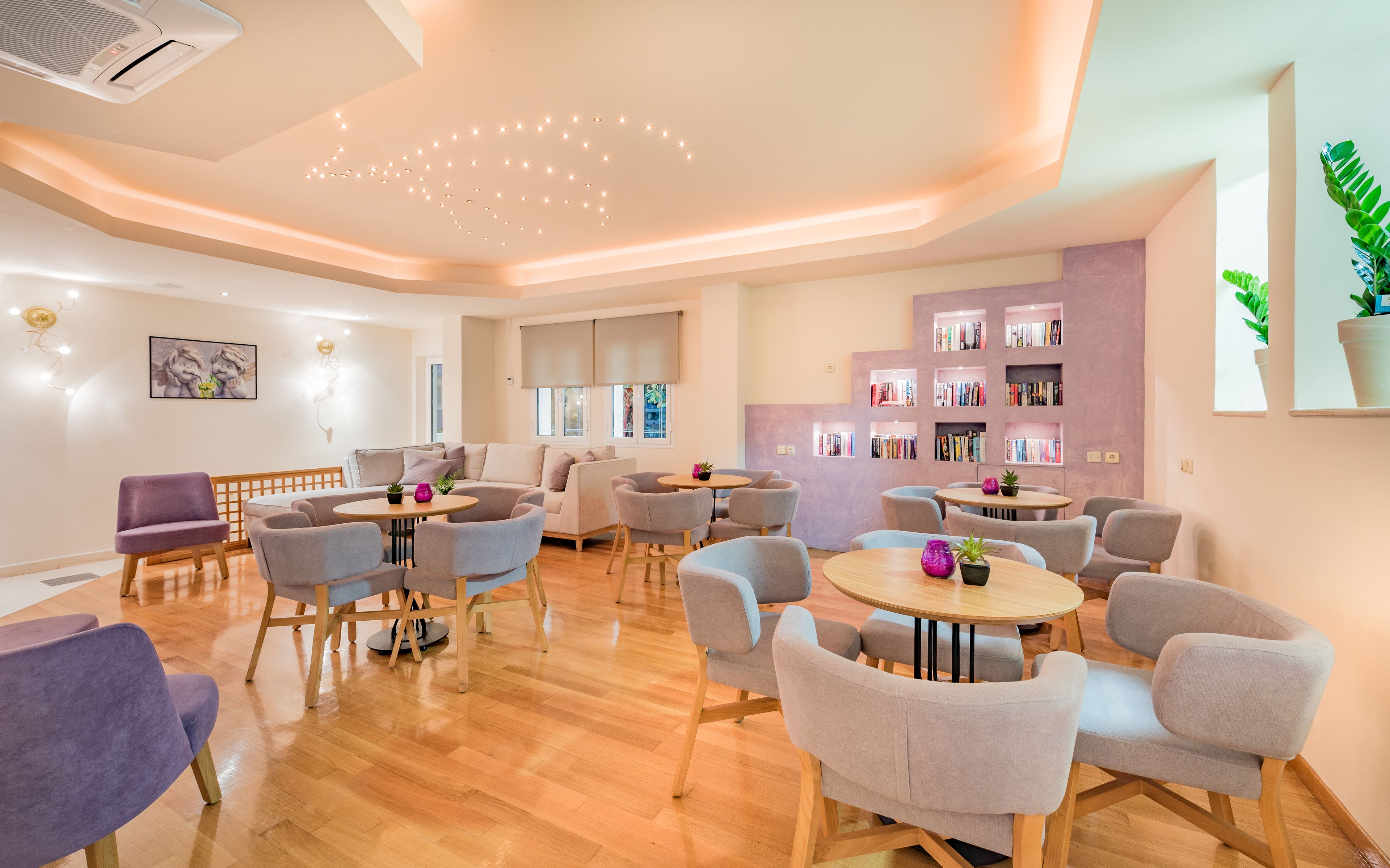 A modern, well-lit room with a wooden floor, multiple seating areas including sofas and chairs, a bookshelf, and a ceiling with recessed lighting.
