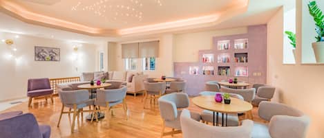 A modern, well-lit room with a wooden floor, multiple seating areas including sofas and chairs, a bookshelf, and a ceiling with recessed lighting.