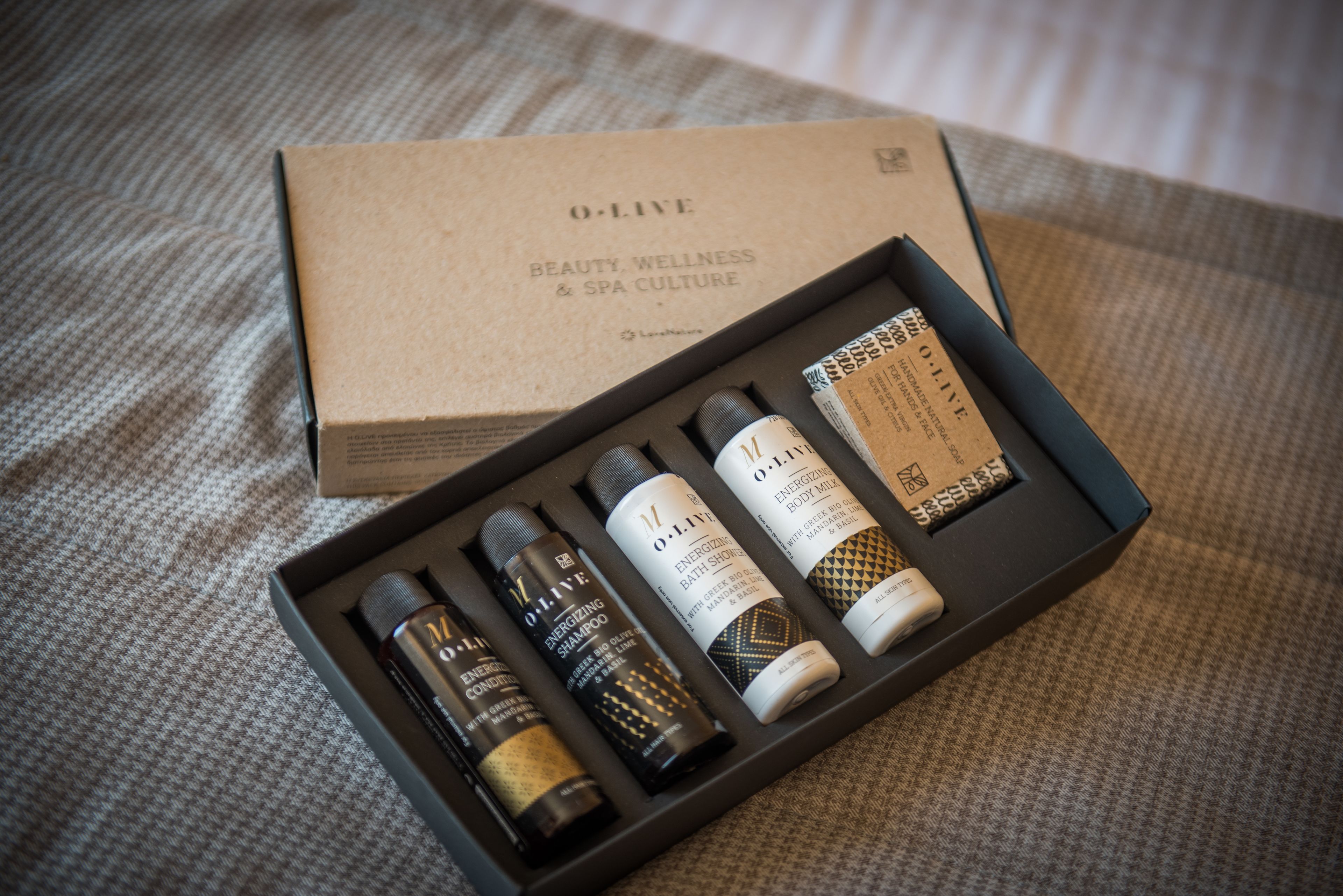 A box of O:Live Beauty, Wellness & Spa Culture products including shampoo, conditioner, body wash, and body scrub.