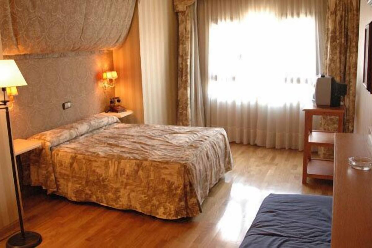 Een hotelkamer met een bed, een bedlampje, een klein tafeltje en een raam met gordijnen.