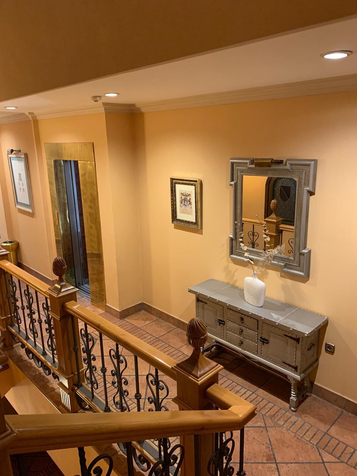 A hallway with a wooden railing, a mirrored wall, and a vintage-style console table with a vase.