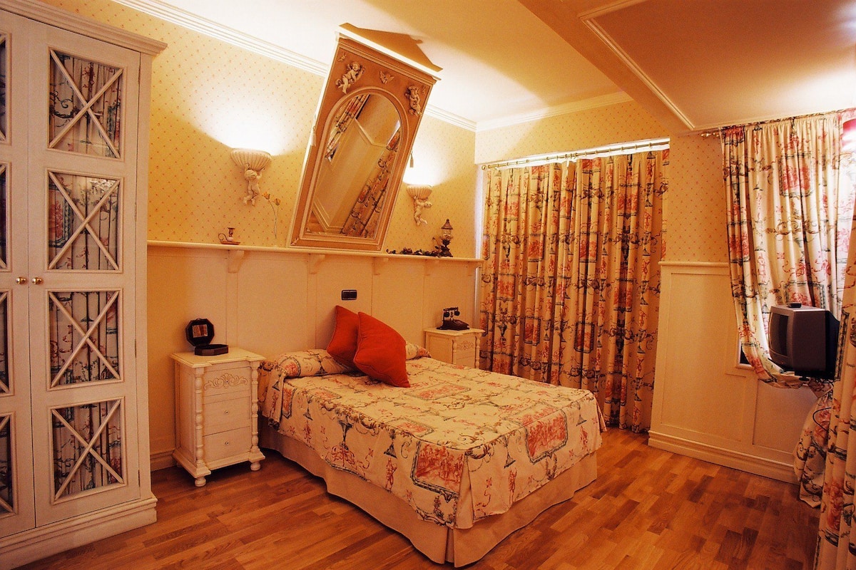 Een slaapkamer met een bed, een dressoir, een nachtkastje, een spiegel, een televisie en gordijnen.