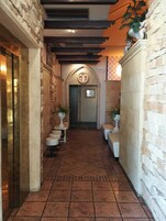 Un pasillo estrecho con paredes de piedra, suelo de baldosas y una puerta al final. Hay plantas en macetas a lo largo de las paredes y un banco con cojín.
