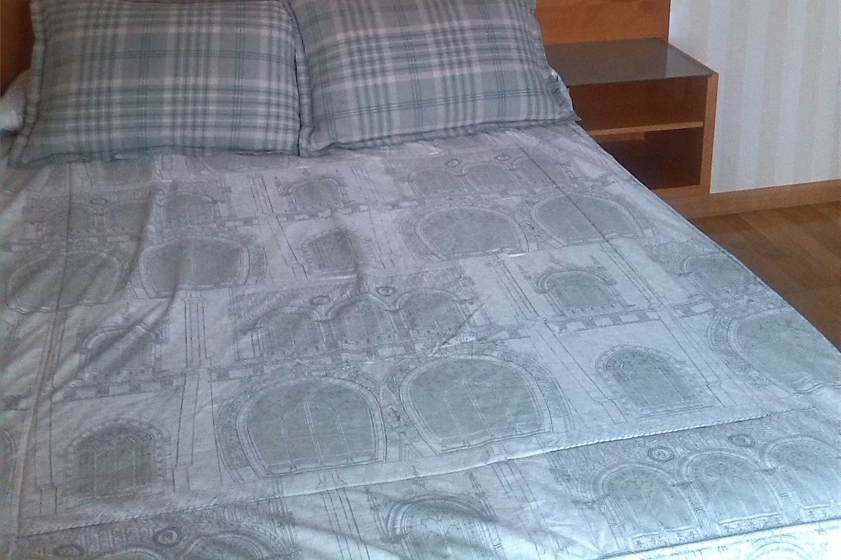 Een bed met een bedsprei met patroon, twee kussens, een houten hoofdbord, een nachtkastje en een wandlamp.