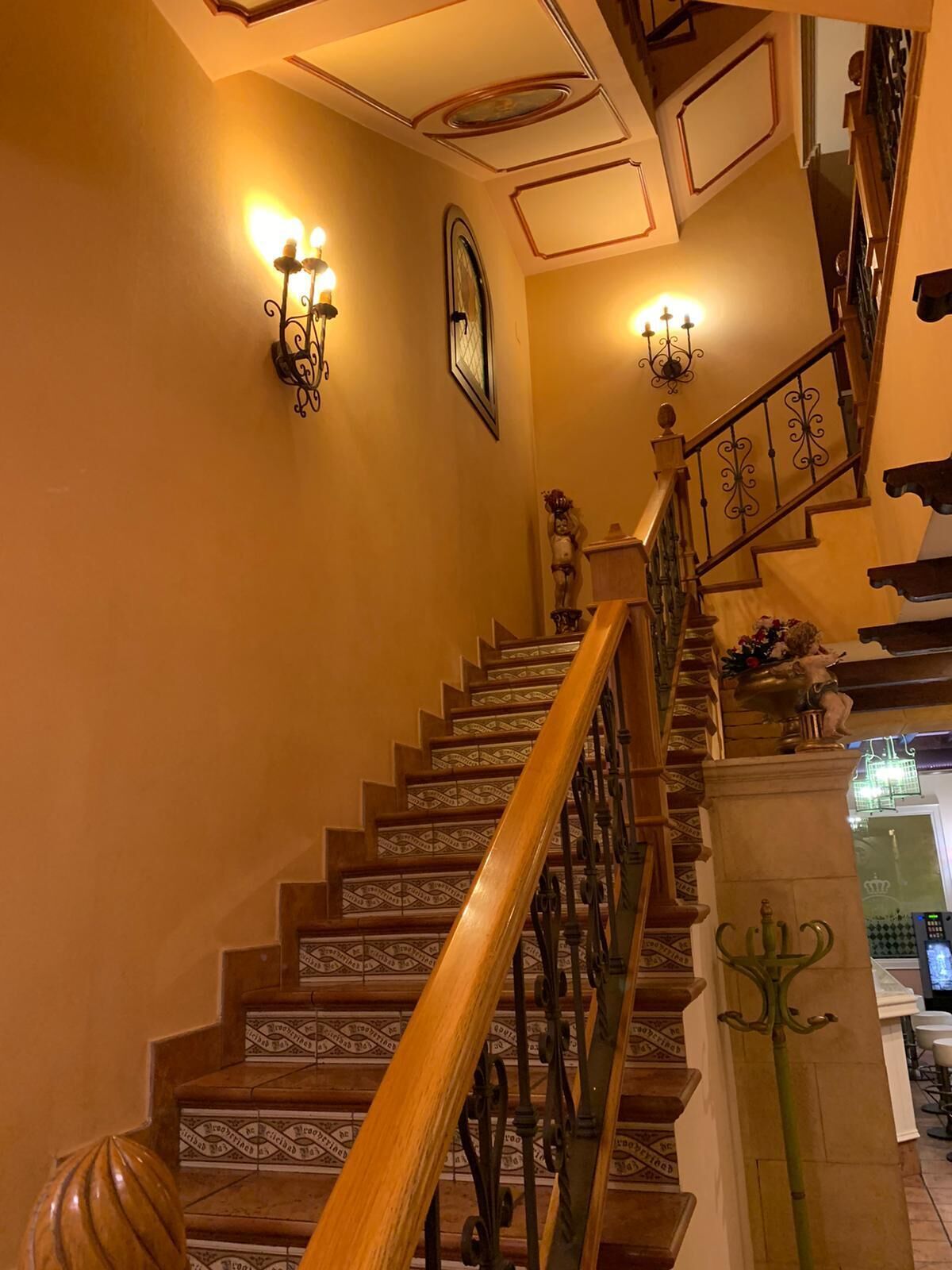 Una escalera con barandillas ornamentales y apliques en la pared, que conduce a un rellano con una urna decorativa y una estatua.
