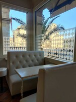 Una zona de estar con un sofá capitonado y una mesita, ventanas amplias con persianas estampadas, y vistas a palmeras y edificios al exterior.