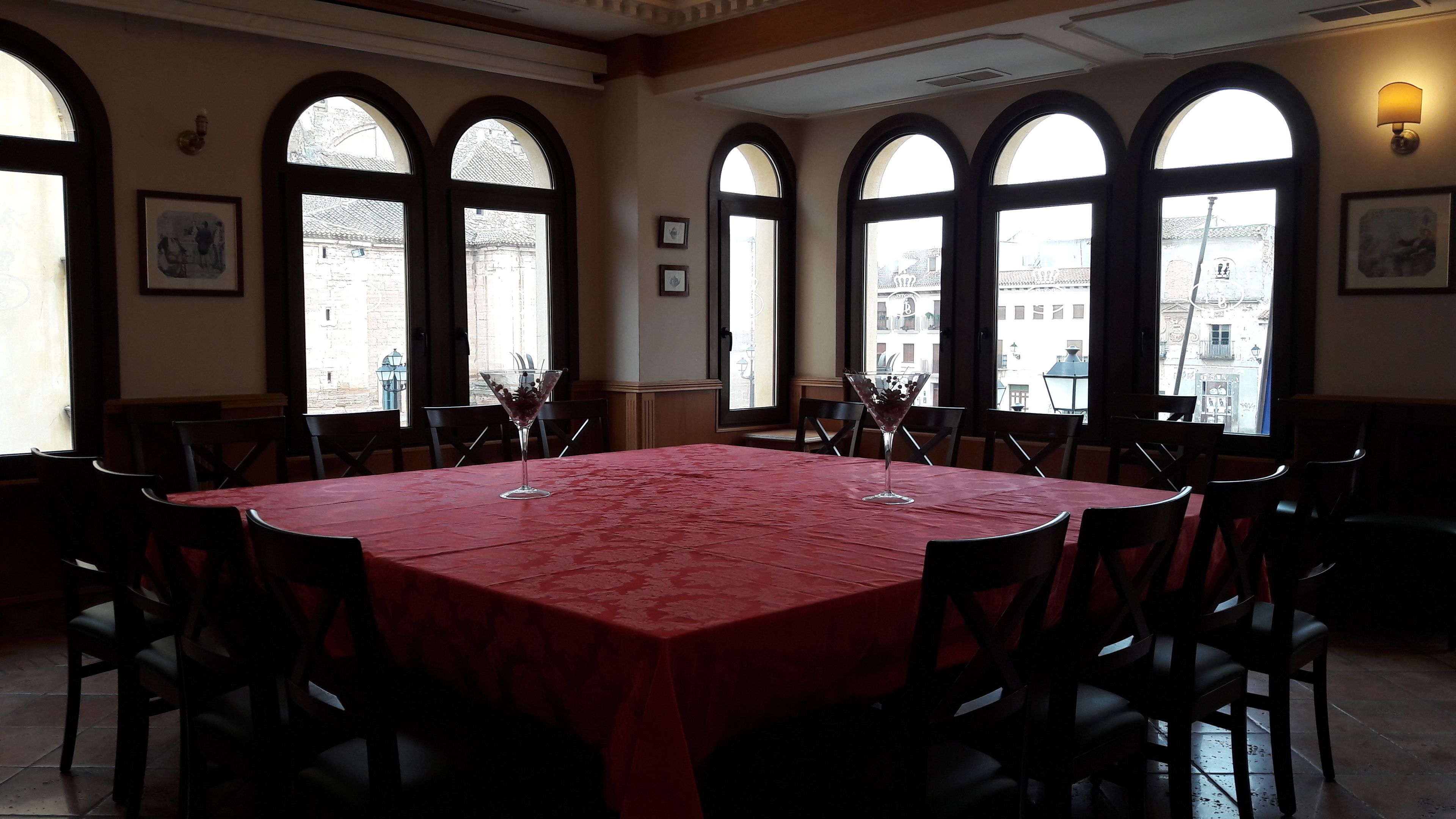 Un comedor con una gran mesa cubierta con mantel rojo, rodeada de sillas y amplias ventanas arqueadas que dan vistas a edificios.