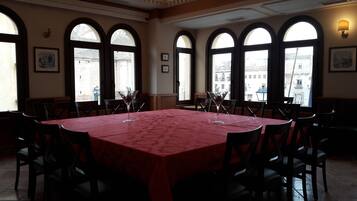 Un comedor con una gran mesa cubierta con mantel rojo, rodeada de sillas y amplias ventanas arqueadas que dan vistas a edificios.