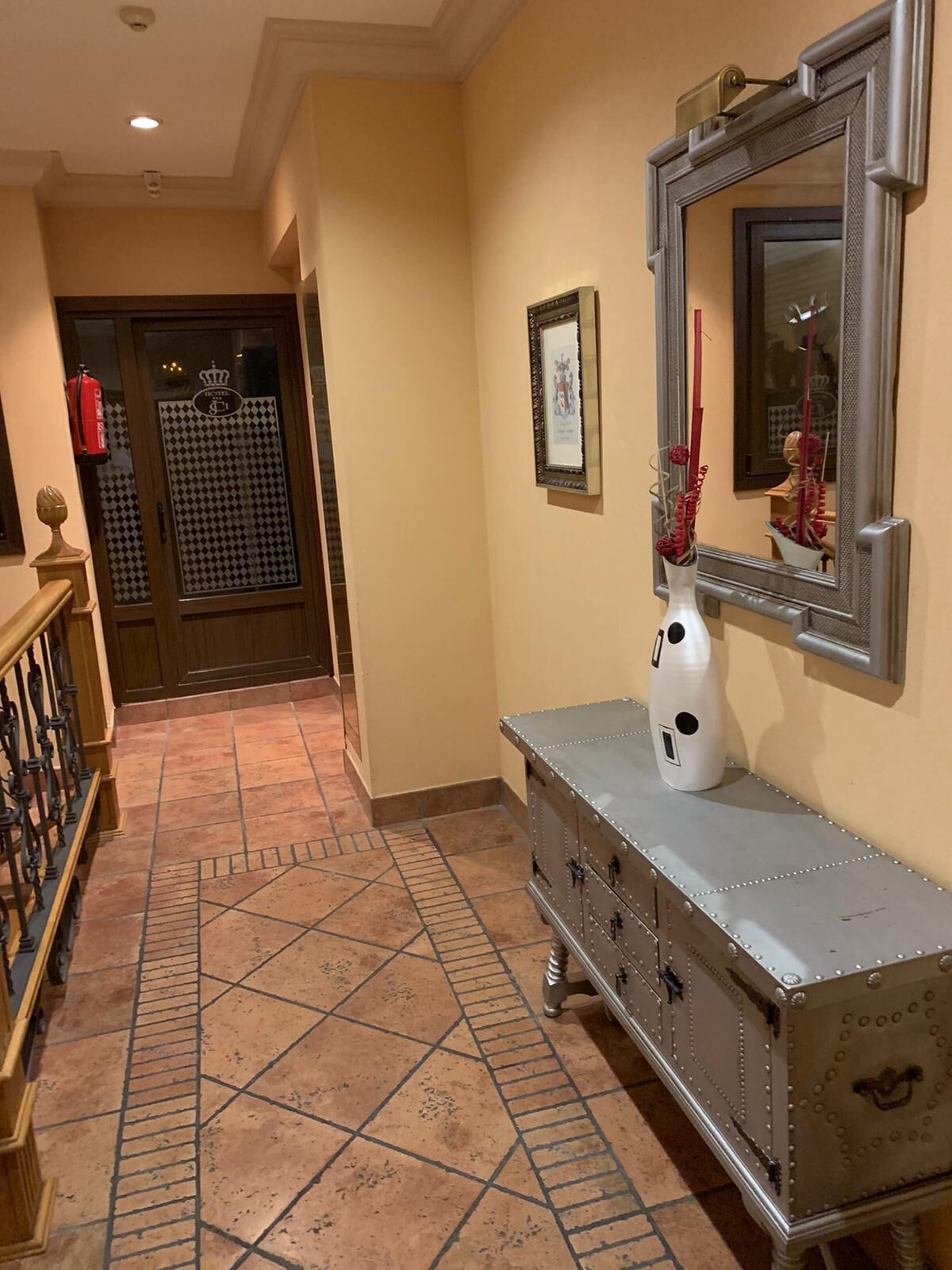 Un corridoio con pavimento piastrellato, ringhiera in legno, un settimino antico, uno specchio con cornice e una porta con pannello di vetro decorativo.