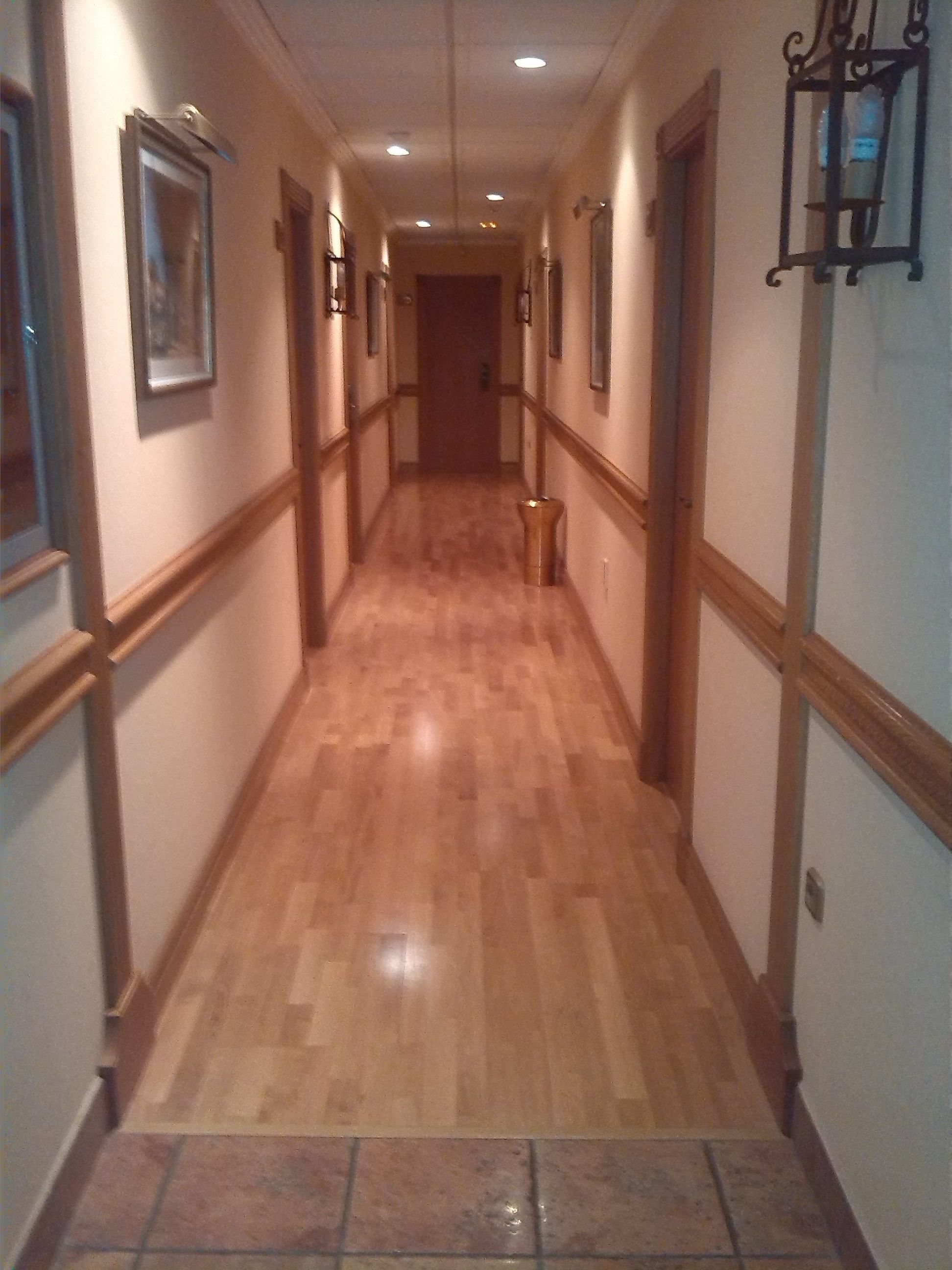 A hallway with wooden flooring and white walls, featuring framed pictures on the left and a wall-mounted light fixture on the right.