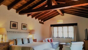 A bedroom with a large bed, a sofa, a small table, and a TV. There are two lamps on nightstands, a window with curtains, and wooden beams on the ceiling. - Areté (Tepoztlán)