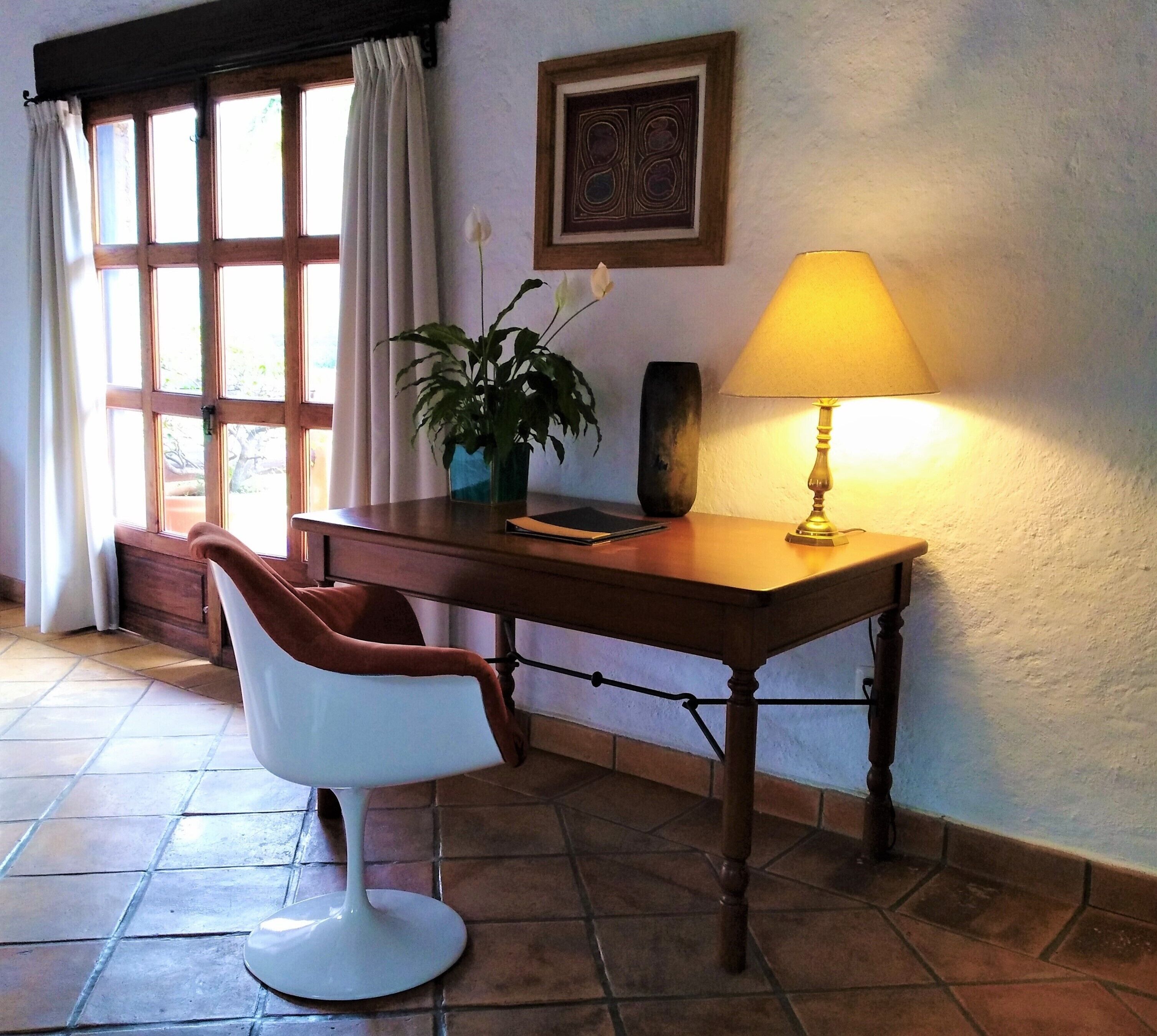 A room with a desk, chair, lamp, potted plant, and a window with curtains.