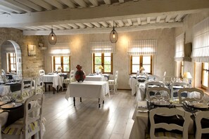 A dining area with white tables and chairs, wooden beams on the ceiling, and large windows with curtains. - Hotel les Hospitaliers (Le Poet-Laval)
