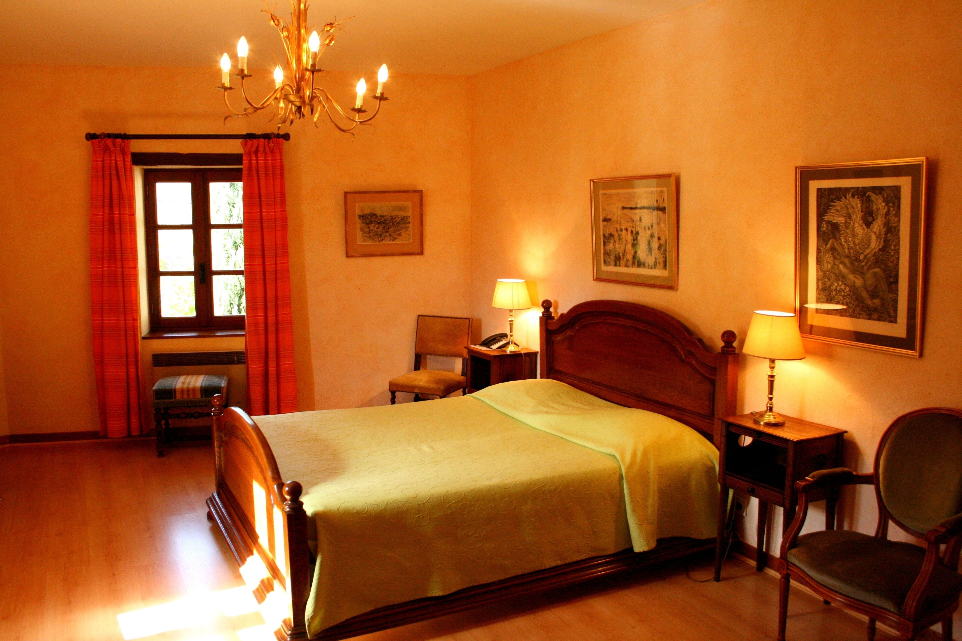 A hotel room with a wooden bed, a chair, a desk with a lamp, and a window with curtains.