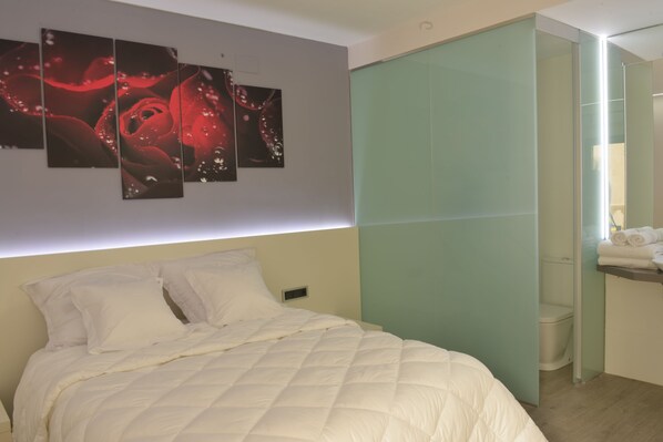 A modern bedroom with a large bed, a nightstand, a wall-mounted light, and a bathroom visible through a glass partition.