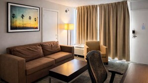 Une chambre d’hôtel avec un canapé marron, une petite table, une chaise, une lampe et un tableau représentant des palmiers.