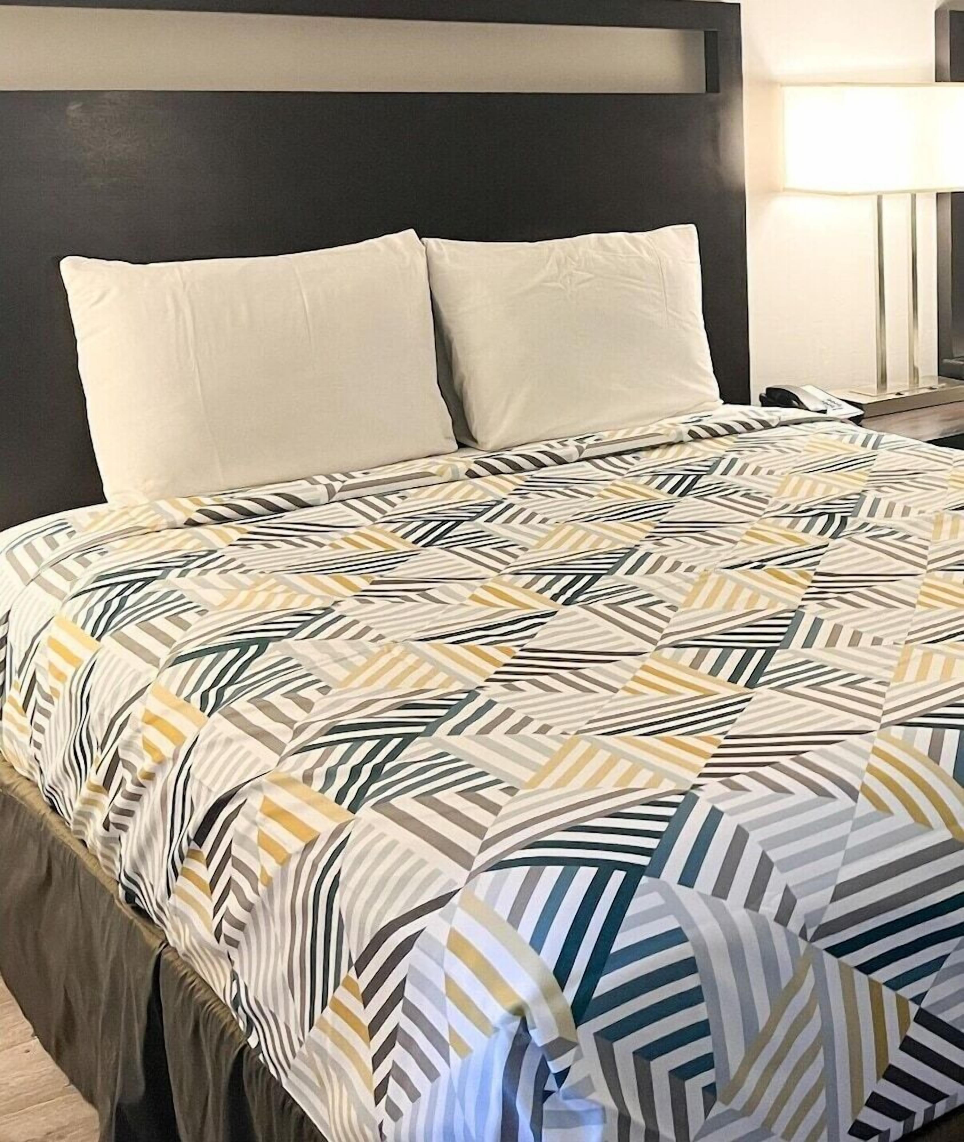 Una cama bien tendida con una colcha estampada, cabecero oscuro y una lámpara de noche.