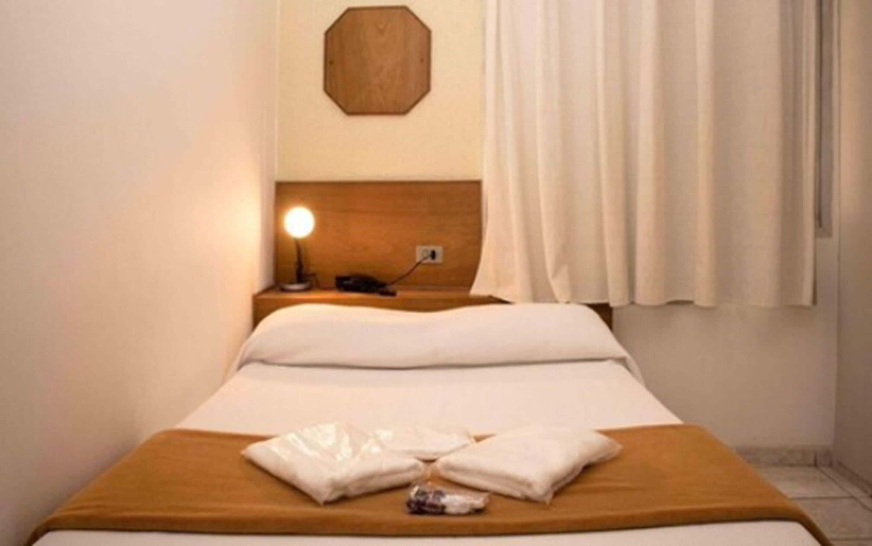 Quarto de hotel com uma cama, mesa de cabeceira com abajur e cabeceira de madeira.