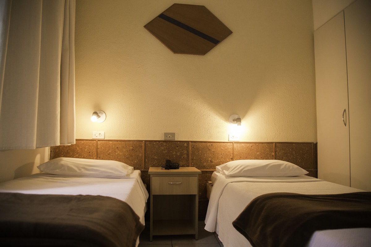 A hotel room with two beds, a nightstand, a small table, and a wardrobe.