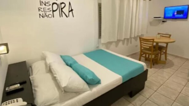 Un dormitorio con cama, mesita de noche y escritorio con silla. Hay un televisor en la pared y un equipo de aire acondicionado sobre el escritorio.
