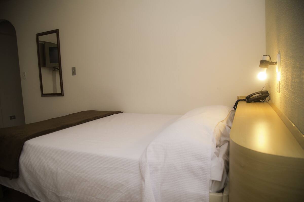 A single bed with a white sheet, a bedside lamp, and a telephone on the nightstand.
