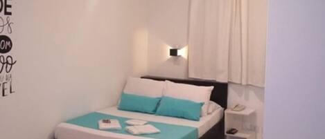 Um quarto com uma cama, mesa de cabeceira, luz na parede e janela com cortinas.