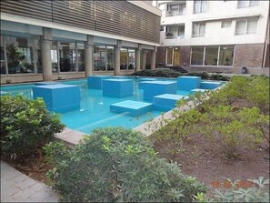A swimming pool with blue rectangular steps, surrounded by greenery and a building with large windows and a brick facade.