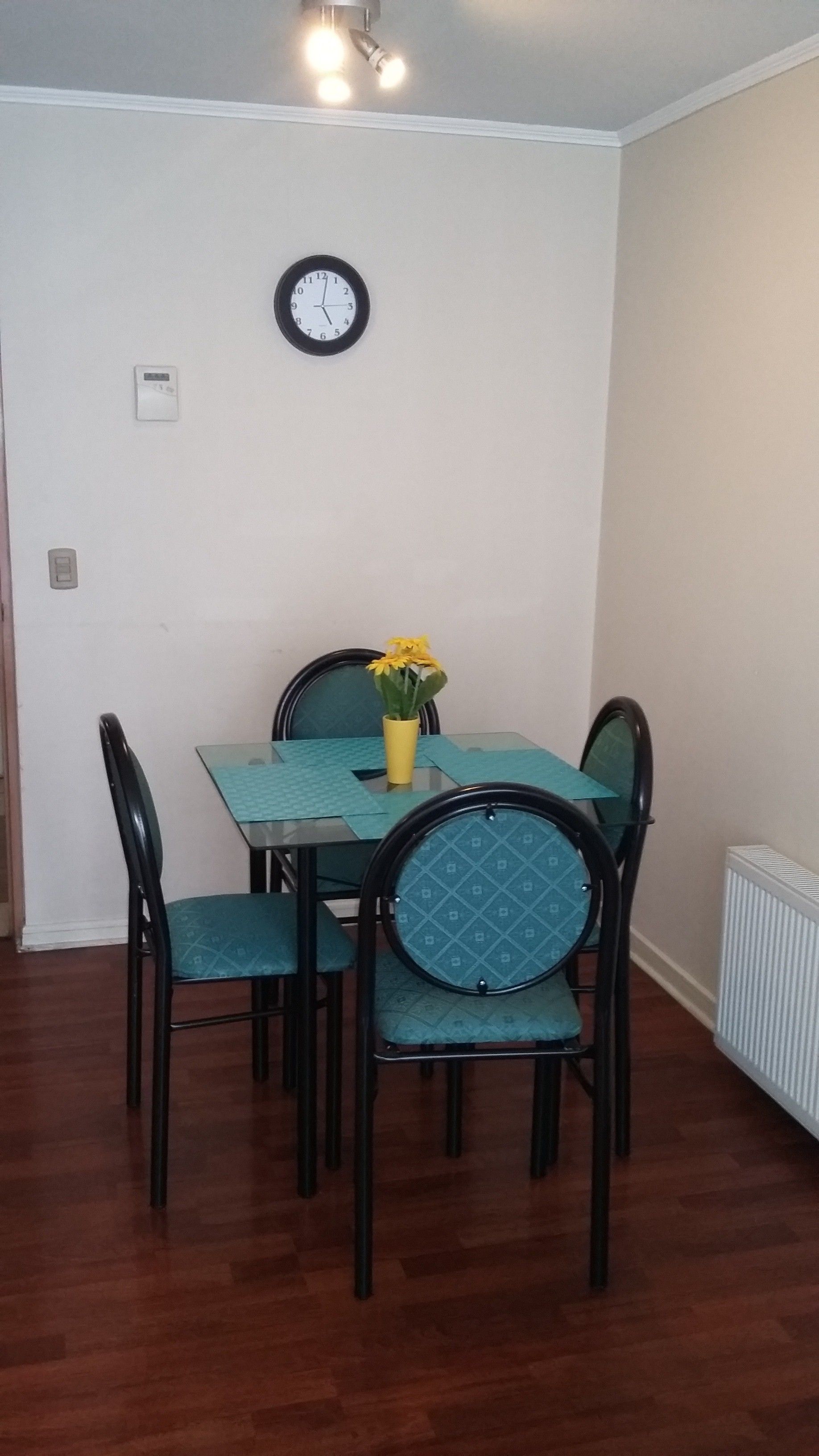 A dining area with a table and four chairs, a clock on the wall, and a radiator on the floor.