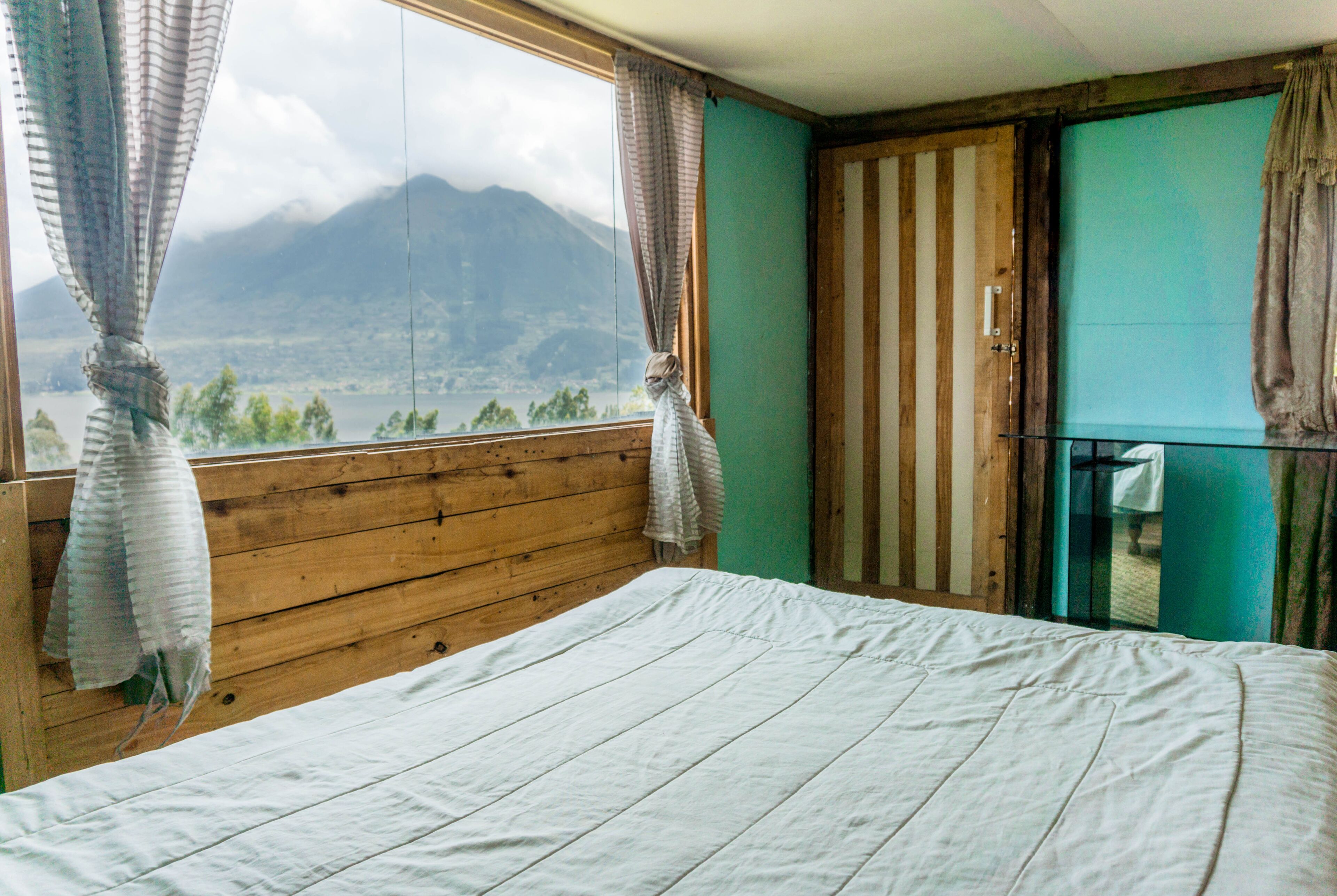 Quarto com cabeceira de madeira, cama com lençol branco, vista para montanhas pela janela e porta com espelho.