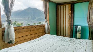 Quarto com cabeceira de madeira, cama com lençol branco, vista para montanhas pela janela e porta com espelho.