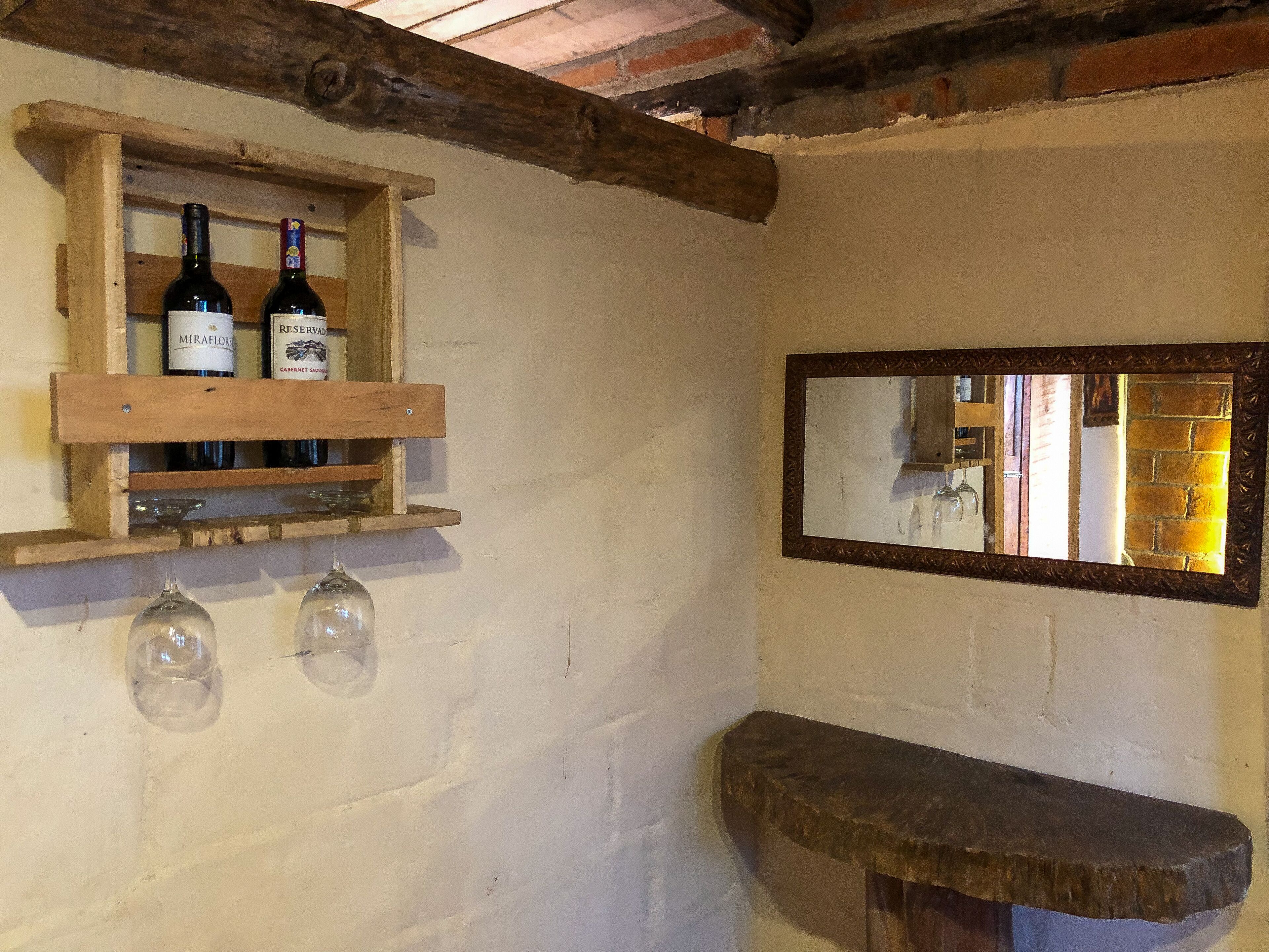 Interior rústico com uma estante de madeira para vinhos com duas garrafas, um espelho e um banco de pedra.