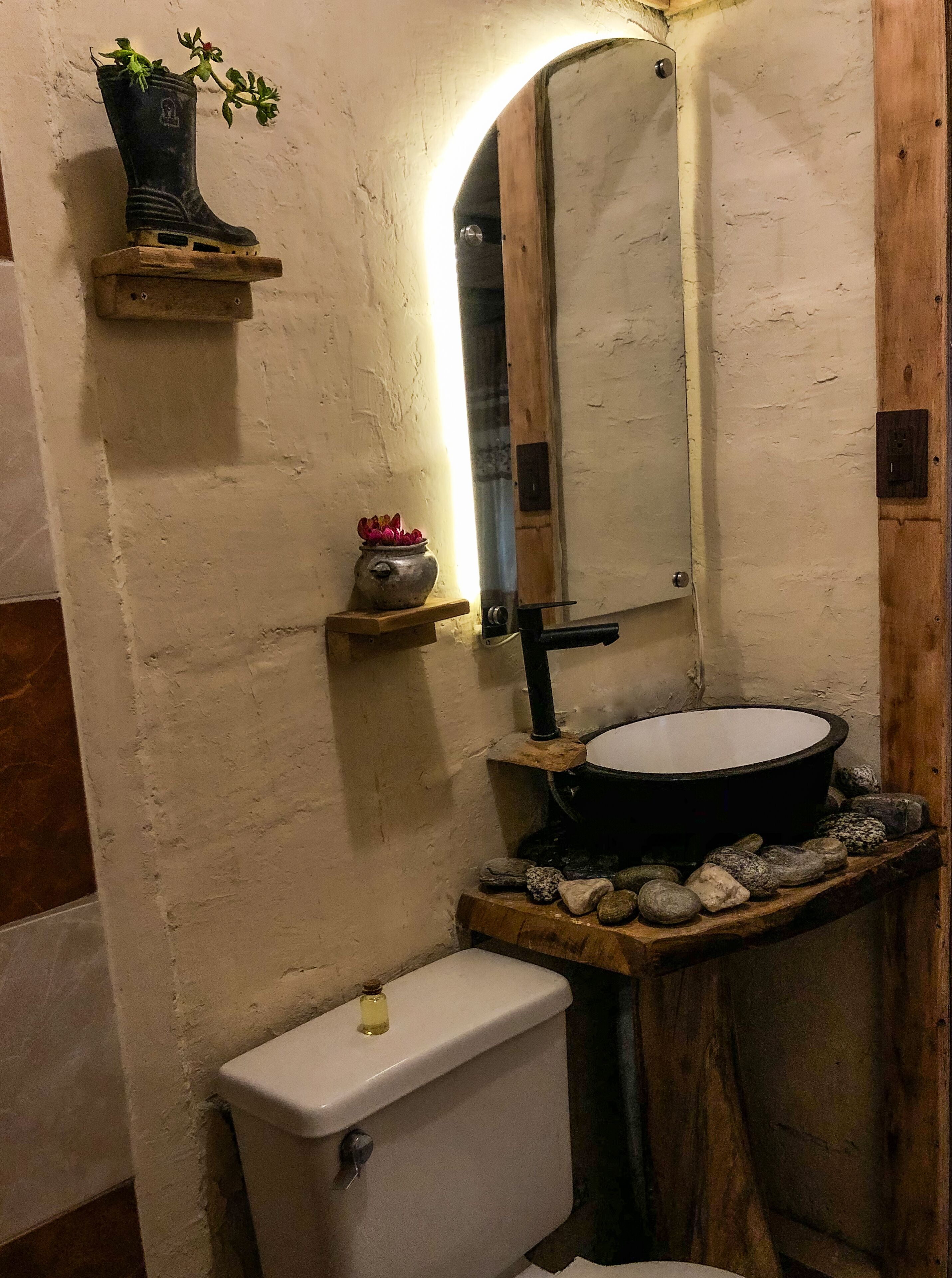 A rustic bathroom with a white toilet, a black basin, and a mirror.