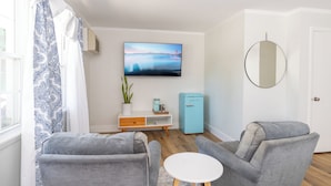 A modern living room with two grey armchairs, a small white table, a blue vintage-style refrigerator, a mounted flat-screen TV, a potted plant, and a large mirror on the wall. - Iron Masters - Inn on Main (Lakeville)