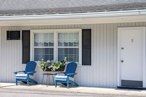 Une maison à l’extérieur blanc, avec une porte d’entrée portant le numéro 5, et deux chaises Adirondack bleues avec des coussins, placées à l’extérieur.