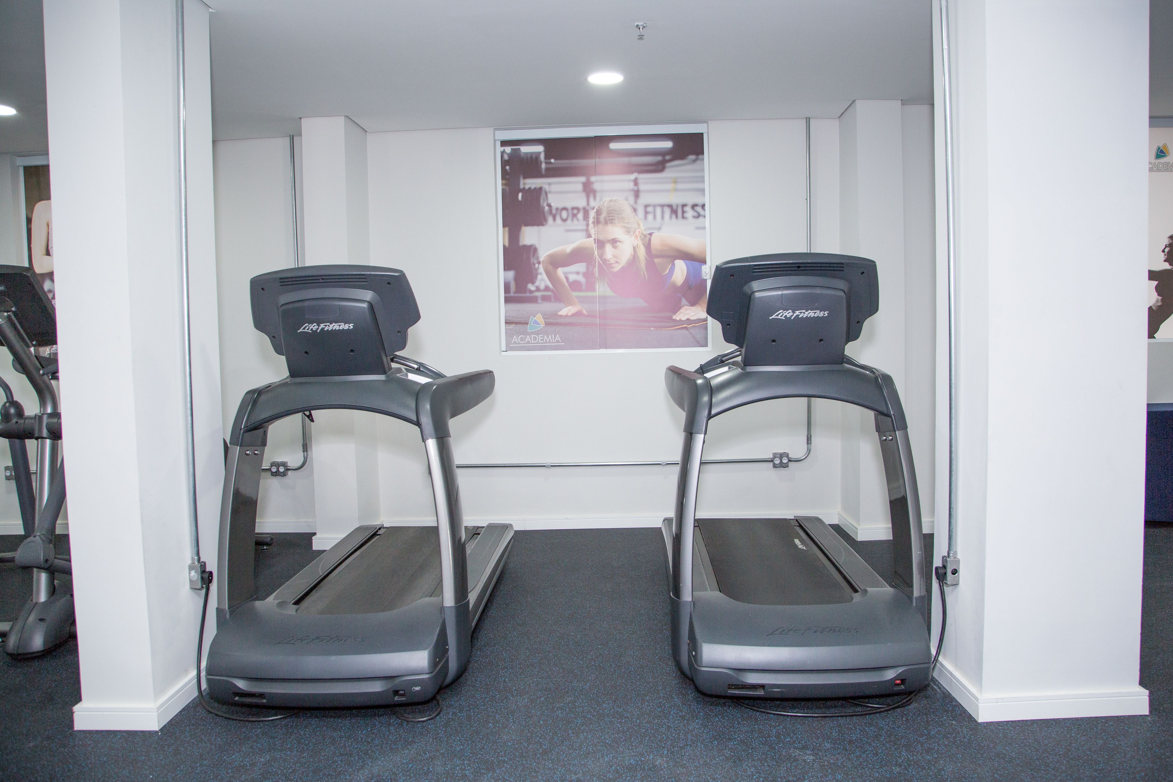 Une salle de sport avec deux tapis de course, une affiche accrochée au mur et un autre tapis de course à l’arrière-plan.