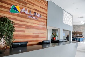 A modern hotel reception area with a wooden wall, a Dell laptop on the counter, and a visible kitchen area in the background.