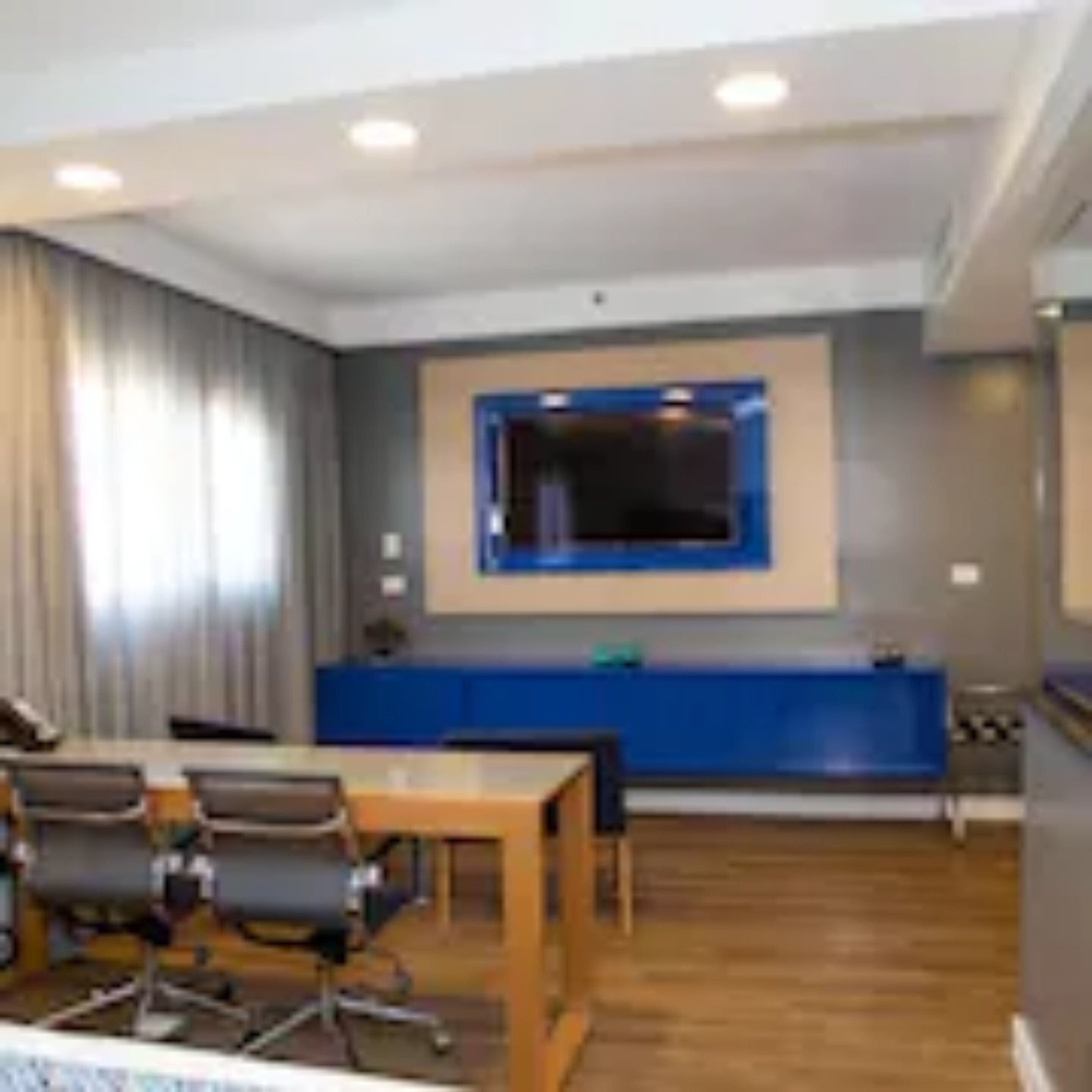 Een moderne hotelkamer met een houten vloer, een bureau met stoelen, een groot raam met gordijnen en een flatscreen televisie die aan de muur hangt.