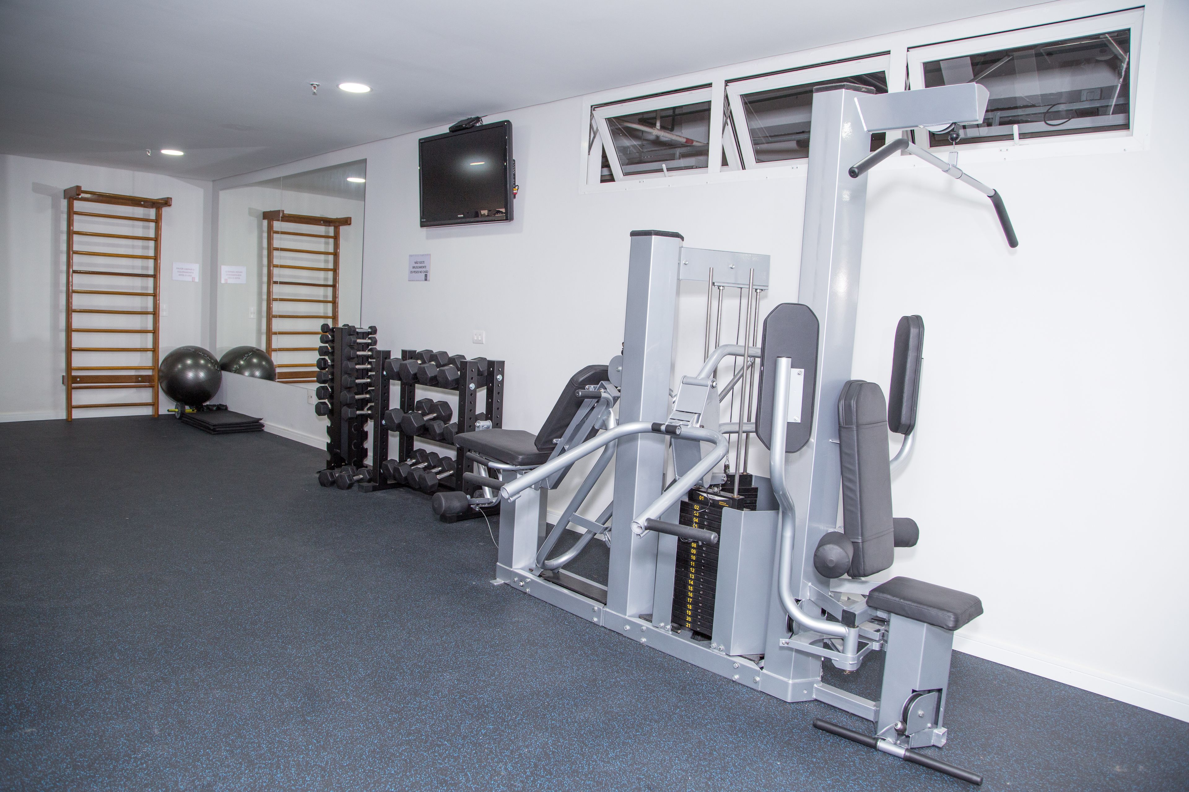 Ett gym med träningsutrustning, en väggmonterad TV och ett fönster med persienner.