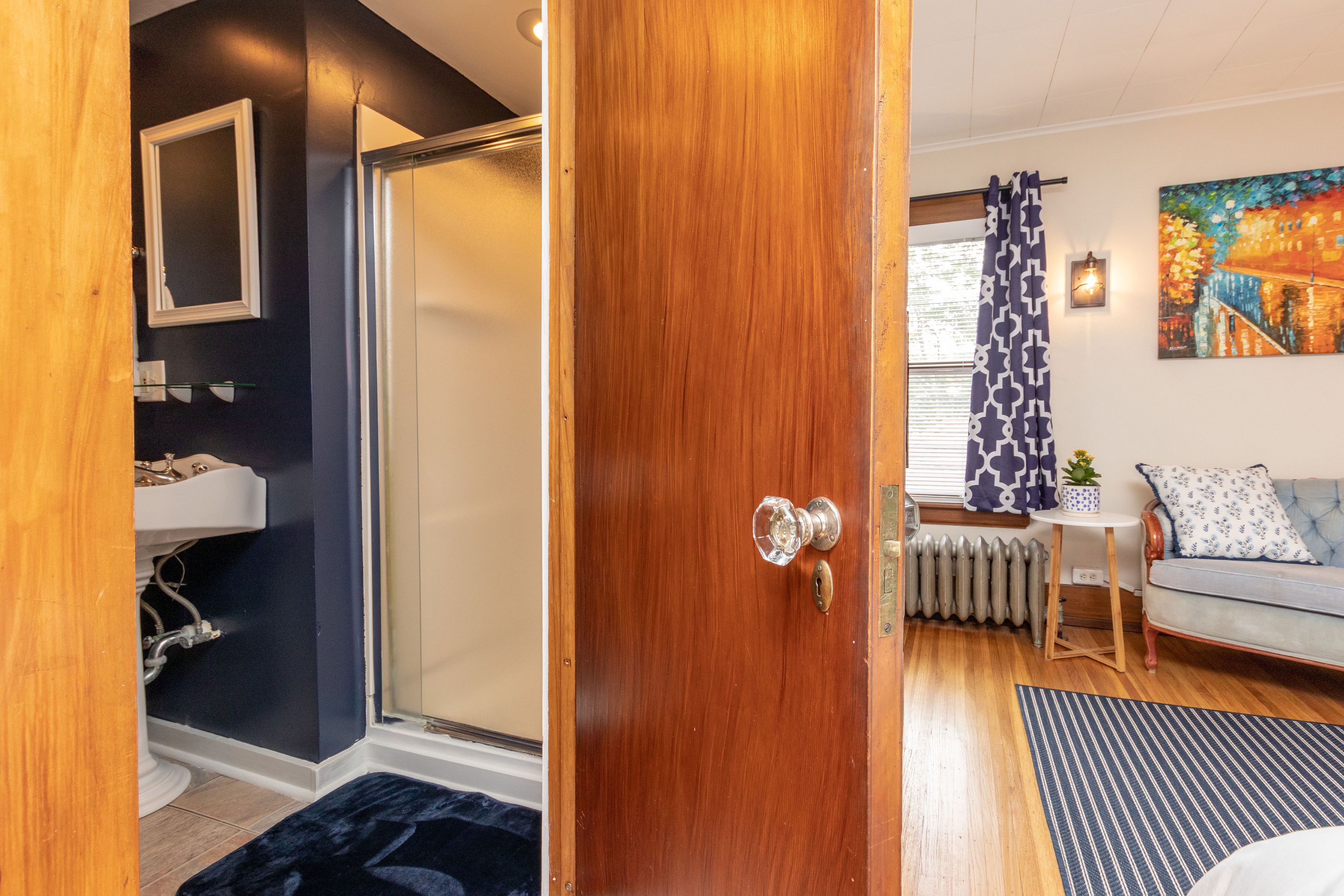 Een kamer met een houten deur, een badkamer met een wit wastafel, een radiator, een bank met geborduurde kussens, en een schilderij aan de muur.