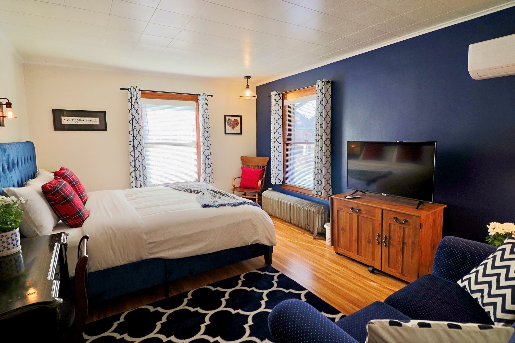 A bedroom with a bed, a TV, a wooden cabinet, a chair, and a window with patterned curtains.