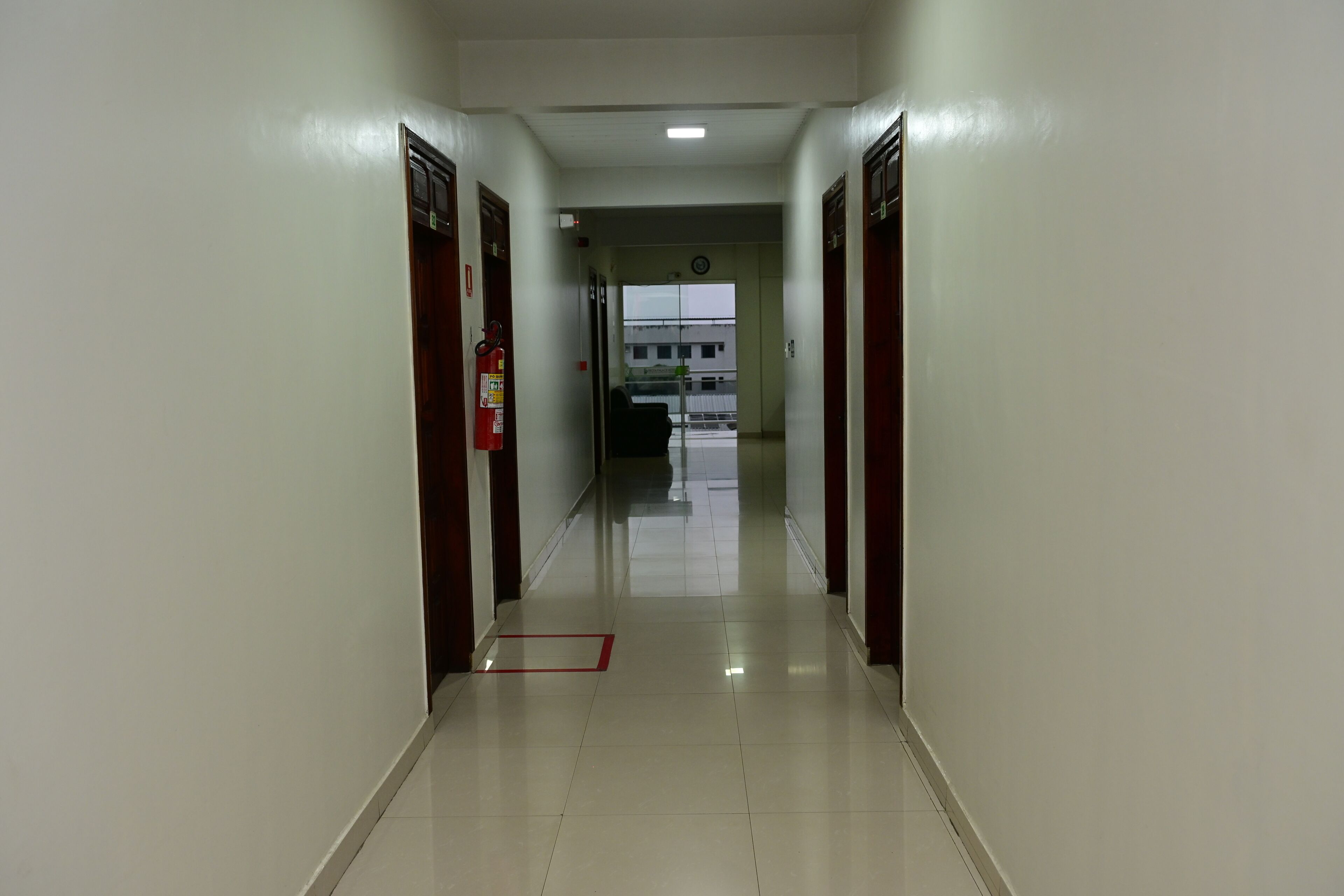 Um corredor com portas em ambos os lados, um extintor de incêndio à esquerda e um relógio na parede.